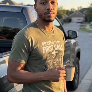 Puede incluir: Una camiseta de manga corta color verde oliva con el texto blanco PRAYER IS MY FIRST CALL sobre una ilustraci&oacute;n gr&aacute;fica de una radio antigua y una cruz. La camiseta se lleva con un collar de cadena de plata fina y vaqueros azules.