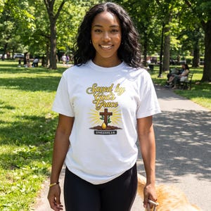 Puede incluir: Una camiseta blanca de manga corta con el texto "Saved by God's Grace" en letra cursiva amarilla y marr&oacute;n, una cruz marr&oacute;n, un peque&ntilde;o brote verde saliendo de tierra marr&oacute;n y el texto "EPHESIANS 2:8" en un recuadro negro.