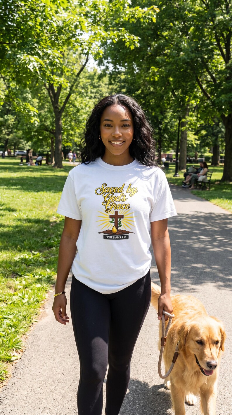 Puede incluir: Una camiseta blanca de manga corta con el texto "Saved by God's Grace" en letra cursiva amarilla y marr&oacute;n, una cruz marr&oacute;n, un peque&ntilde;o brote verde saliendo de tierra marr&oacute;n y el texto "EPHESIANS 2:8" en un recuadro negro.