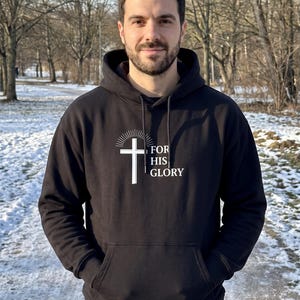 May include: A black hooded sweatshirt featuring a white graphic of a cross with radiating lines above it and the text FOR HIS GLORY printed to the right. The garment includes a front kangaroo pocket, long sleeves, and a drawstring hood.