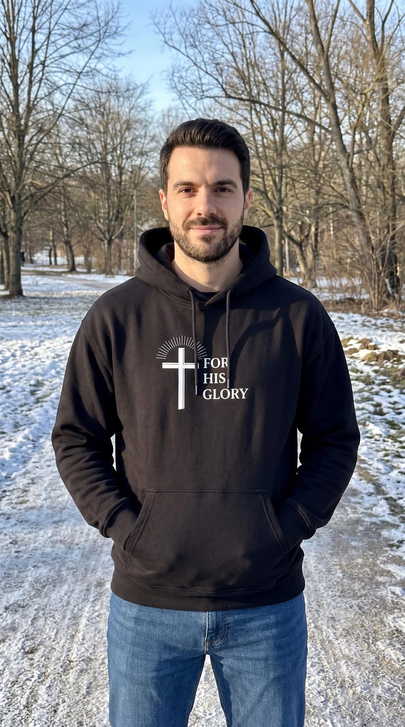 May include: A black hooded sweatshirt featuring a white graphic of a cross with radiating lines above it and the text FOR HIS GLORY printed to the right. The garment includes a front kangaroo pocket, long sleeves, and a drawstring hood.