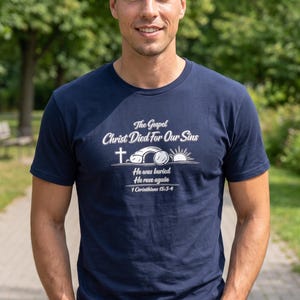 Puede incluir: Una camiseta azul marino de manga corta con texto en blanco que dice The Gospel Christ Died For Our Sins, He was buried, He rose again, 1 Corinthians 15:3-4, acompañada de un gráfico de una cruz, una tumba abierta y un sol naciente.