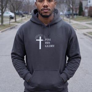 May include: A charcoal gray hooded sweatshirt featuring a white graphic of a cross with radiating lines above it and the text FOR HIS GLORY printed to the right. The hoodie includes a front kangaroo pocket and matching drawstring cords.
