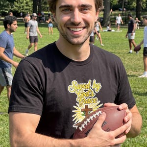 Puede incluir: Una camiseta negra de manga corta con un estampado gr&aacute;fico que incluye el texto "Saved by God's Grace" en letras cursivas amarillas sobre una cruz marr&oacute;n y un dise&ntilde;o de rayos de sol amarillos, sostenida junto a un bal&oacute;n de f&uacute;tbol americano de cuero marr&oacute;n.
