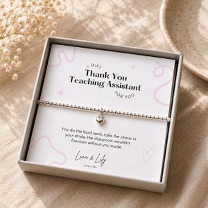 Könnte beinhalten: Ein silbernes Perlenarmband mit einem kleinen Herzanhänger in einer Geschenkbox. Die Präsentationskarte enthält den Text "A Gift Thank You Teaching Assistant For You" und "You do the hard work, take the chaos in your stride, the classroom wouldn't function without you inside" sowie "Luna & Lily Jewellery" am unteren Rand.