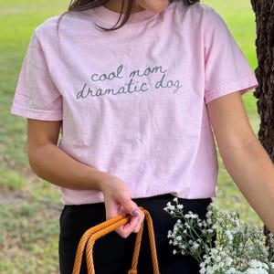 May include: A light pink short-sleeved t-shirt featuring the dark gray text "cool mom dramatic dog" printed in a cursive font across the chest. The garment has a crew neckline and a standard fit, providing a simple and casual appearance.