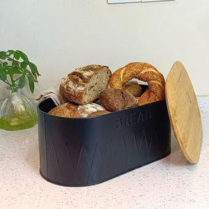May include: A black bread box with a wooden lid, filled with various types of bread. The box has a diamond pattern and the word "BREAD" is embossed on the front. The bread includes a loaf, bagels, and rolls.