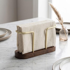 May include: A wooden napkin holder with a dark brown base and a gold-coloured metal frame. It holds a stack of beige cloth napkins. The setting is a well-lit table with a marble surface.