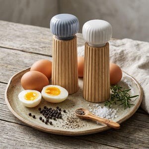 Puede incluir: Dos molinillos de sal y pimienta de madera con diseño acanalado, rematados con cerámica azul y blanca. Los molinillos están sobre un plato con huevos, granos de pimienta, sal y una pequeña cuchara de madera. Tema culinario.
