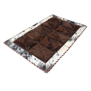 May include: A rectangular rug constructed from stitched panels of animal hide. The central section features solid dark brown fur, while the border consists of white hide with black and grey speckled patterns, finished with a whip-stitched edge.