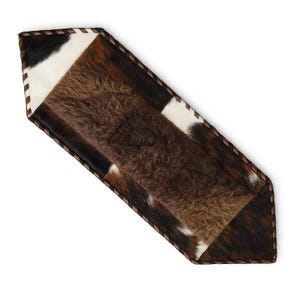 May include: A hexagonal table runner featuring a central panel of textured brown sheepskin surrounded by panels of cowhide in shades of black, white, and brown. The entire piece is finished with a dark brown stitched leather edge trim.