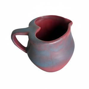 May include: A handmade ceramic pitcher with a unique heart-shaped opening. The pitcher features a handle and a spout. The exterior has a blend of red and blue-gray hues, creating a marbled effect. The interior is a solid red color.