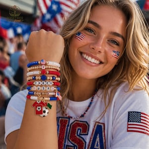 May include: A collection of red, white, and blue beaded bracelets with gold details and charms. Some bracelets spell out "CHEER USA". One charm is a soccer jersey with a soccer ball. The bracelets are worn on a wrist.