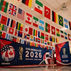 Op de afbeelding: Een presentatie voor het FIFA Wereldkampioenschap 2026, met een banner met de tekst "FIFA WORLD CUP 2026 UNITED STATES CANADA MEXICO" en een voetbalontwerp. Nationale vlaggen hangen aan het plafond, met de hashtag "#WEARE26".