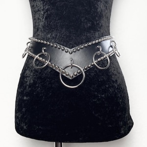 May include: A black V-shaped belt featuring silver-toned metal hardware, including a series of small spherical studs along the top and bottom edges, and five large circular rings attached to the front and sides of the leather-like material.