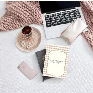 May include: A flat lay featuring a laptop, journal, phone, and accessories on a white surface. A pink knitted blanket and a small plate with a drink and earrings are also present. The journal has a beige and white checkered pattern.