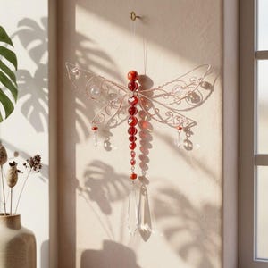 May include: A decorative dragonfly made of copper wire, red beads, and clear crystal accents. The dragonfly is hanging on a wall. The wings have intricate wire detailing and the body is made of graduated red beads. Clear crystal pendants hang from the wings and tail.