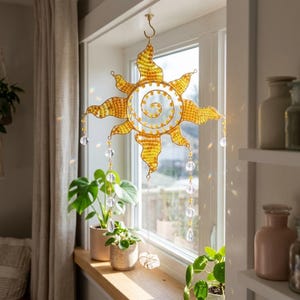 May include: A golden suncatcher with a spiral center and crystal accents hangs in front of a window. The sun-shaped decoration has a textured, woven appearance and is suspended by a hook. Clear crystals dangle from the sun's rays, adding sparkle.