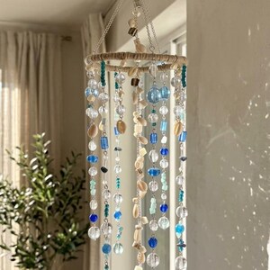 Puede incluir: Móvil colgante con un marco circular envuelto en hilo natural, adornado con hebras de cuentas transparentes, azules y verdes, junto con conchas marinas. Suspendido de un gancho plateado.