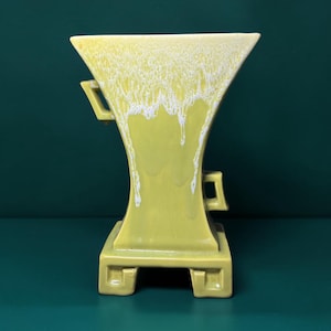 May include: A ceramic vase with a flared top and a square base, featuring a glossy chartreuse yellow glaze with a white drip pattern cascading from the rim. The vessel includes two small, angular handles on opposite sides of the body.