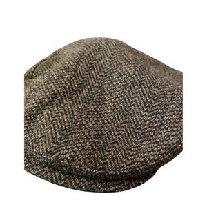May include: A flat cap featuring a brown and black herringbone pattern. The cap has a rounded crown and a short, stiff brim at the front. The fabric displays a textured, woven appearance with a consistent zigzag design throughout the entire surface.