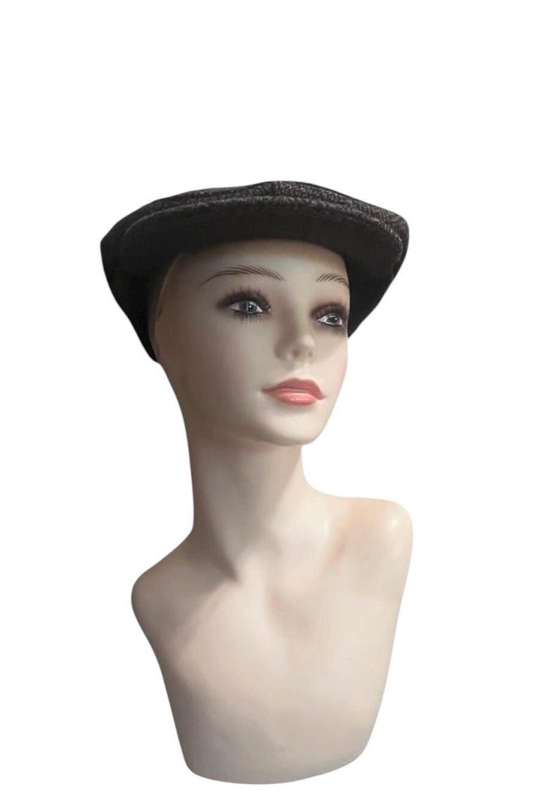 May include: A dark gray and black herringbone patterned flat cap made of a textured wool-like fabric. The cap features a rounded crown and a short, stiff brim that extends over the forehead, providing a classic silhouette and structured shape.
