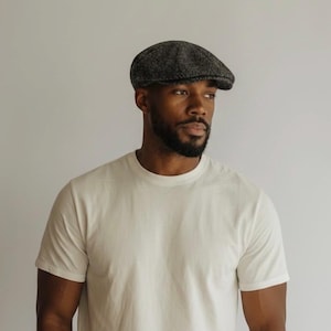 May include: A charcoal grey and black speckled flat cap with a rounded crown and a short, stiff brim. The cap features a textured, woven fabric construction with a classic panel design that sits low on the head, providing a structured silhouette.