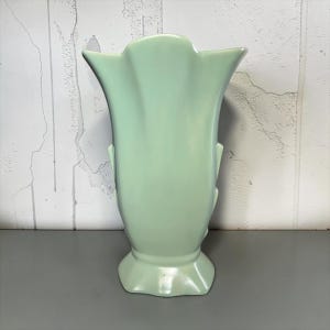 May include: A pale mint green ceramic vase with a smooth, glossy finish. The vase features a flared, scalloped rim, a tapered body with vertical molded ridges along the sides, and a wide, pedestal-style base with a ruffled edge design.
