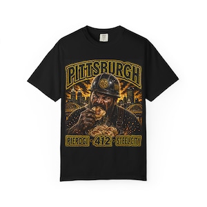 Peut inclure: T-shirt noir avec un motif graphique doré. Le motif comprend le mot "PITTSBURGH" au-dessus d'une illustration d'une personne mangeant des pierogis. Le texte "PIEROGI 412 STEEL CITY" est également inclus.