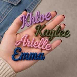 Könnte beinhalten: Vier bestickte Namensaufnäher in Schreibschrift. Der oberste Aufnäher ist lila und zeigt den Namen Khloe, der zweite ist olivgrün und zeigt Kaylee, der dritte ist hellrosa und zeigt Arielle, und der unterste Aufnäher ist dunkelblau und zeigt Emma.