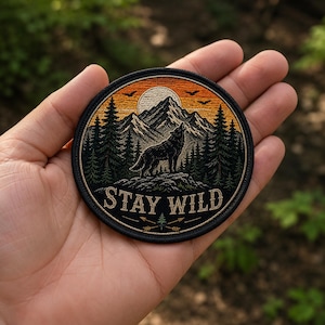 Puede incluir: Un parche bordado circular que muestra un lobo negro aullando a una luna blanca sobre un cielo naranja al atardecer. El diseño incluye pinos de color verde oscuro, montañas escarpadas y el texto STAY WILD en fuente serif sobre dos flechas cruzadas.