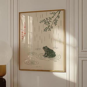 以下が含まれることがあります： 雨と葉のある池にいる緑色のカエルを描いた額入りのアートプリント。アートワークはベージュの背景と金のフレームです。日本語のテキストがプリントの左側に表示されます。