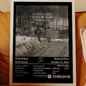 May include: A framed poster featuring the Noah Kahan Stick Season album cover with a snowy road scene, tracklist, release date of October 14, 2022, a total album length of 55:21, a signature, and a Spotify code at the bottom on a black background.