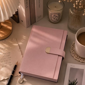 Könnte beinhalten: Ein rosa Lederjournal mit goldenem Verschluss steht im Mittelpunkt dieses Bildes. Ein schwarz-goldener Stift, eine Tasse Kaffee und eine brennende Kerze sind ebenfalls zu sehen. Das Journal ist geschlossen und hat ein Schleifenlesezeichen.
