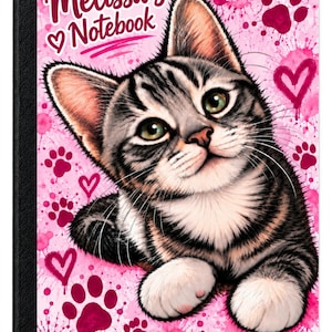 May include: A notebook with a black spine and a front cover featuring a gray tabby kitten on a pink background with paint splatters, paw prints, and hearts. The text "Melissa's Notebook" is printed in white and pink at the top left corner.