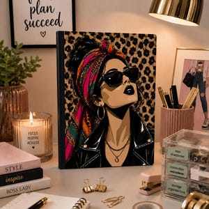 May include: A rectangular notebook featuring a graphic illustration of a person wearing a leopard print headscarf, black sunglasses, and a black leather jacket. The background of the illustration is a repeating leopard print pattern.