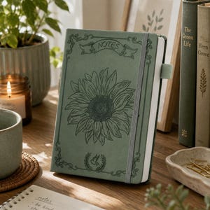 May include: A sage green notebook featuring a sunflower illustration and the word "NOTES". The notebook is secured with an elastic band. A small notepad with handwritten notes and a pen are visible in the foreground.