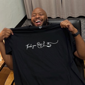May include: A black short-sleeved t-shirt featuring white cursive text that reads Thank you, Ray J, accompanied by a graphic illustration of a hand gesture. The shirt has a crew neckline and is made of a solid black fabric with white ink.