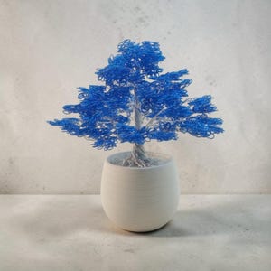 Op de afbeelding: Handgemaakte bonsai boomsculptuur met helderblauwe draadbladeren. De zilverkleurige stam staat in een witte, afgeronde keramische pot. Een uniek decoratief item voor thuis of op kantoor.