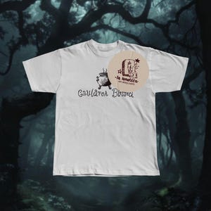 May include: White t-shirt featuring a black illustration of a cauldron and the words "Cauldron Blessed". A circular logo with text and a rabbit is on the right. The shirt is set against a dark, forest backdrop.