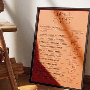 Op de afbeelding: Een ingelijste oranje bibliotheekkaartprint met een zwarte rand. De kaart bevat titels en publicatiejaren van boeken, waaronder "Haunting Adeline". De naam van de lener is "Joanna". De tekst ".la lunoteca." staat rechtsonder.