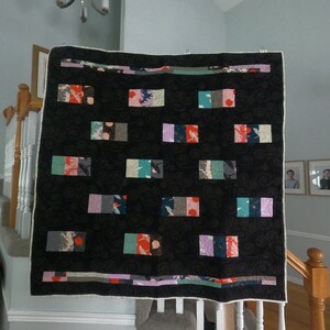 May include: A black and white quilt with a repeating pattern of colorful squares. The quilt is hanging on a white wall with a wooden staircase in the background.