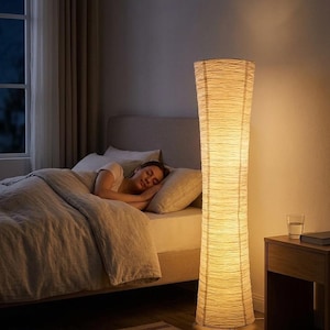 May include: A tall, cylindrical floor lamp with a textured, off-white shade illuminates a bedroom. The lamp stands next to a bed where a person is sleeping. A nightstand with a glass of water and a book is also visible.