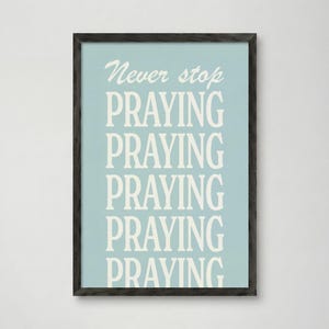 Puede incluir: Una impresión artística enmarcada con un fondo azul claro y las palabras "Never stop PRAYING" en blanco. La palabra "PRAYING" se repite cinco veces. El marco es gris oscuro.