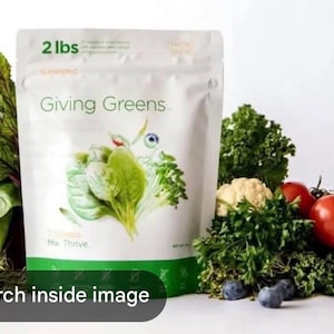 May include: A white bag of "Giving Greens" with a green stripe, containing a green powder, is surrounded by fresh vegetables. The bag is labelled "2 lbs". Fresh spinach, tomatoes, cauliflower, and blueberries are also visible.