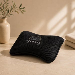 May include: A black rectangular pillow with a textured mesh surface and curved edges. The centre features a white graphic logo of a sunburst above the text HEAD SPA. The pillow has a structured form and a consistent dark colour throughout its design.