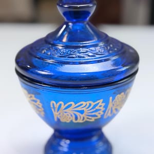 Op de afbeelding: Een kobaltblauwe glazen schaal met bijpassend deksel. De kom en het deksel zijn voorzien van een goudkleurig bloemmotief. De schaal heeft een voetstuk en een knopvormige handgreep op het deksel. Het glas is doorschijnend en diepblauw van kleur.