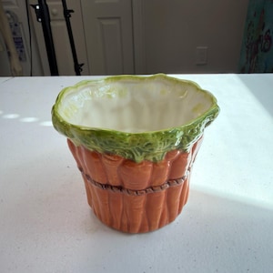 Peut inclure: Un pot de fleurs en céramique en forme de botte de carottes orange liées par une cordelette marron. Le bord supérieur présente un motif de feuilles vertes texturées, tandis que l'intérieur du pot est émaillé d'un blanc cassé lisse.