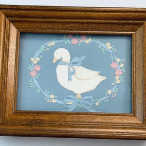 Può includere: Una cornice rettangolare in legno con finitura marrone medio contenente la stampa di un'oca bianca che indossa un fiocco azzurro. L'oca è centrata all'interno di una ghirlanda ovale di piccoli fiori rosa e gialli su uno sfondo blu.