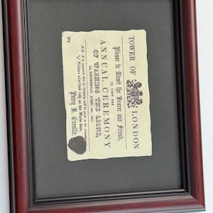 May include: A framed historical document titled Tower of London Annual Ceremony of Washing the Lions, dated Wednesday, April 1st, 1857. The off-white paper features black printed text, a royal crest, and a dark wax seal, set within a dark wood frame.